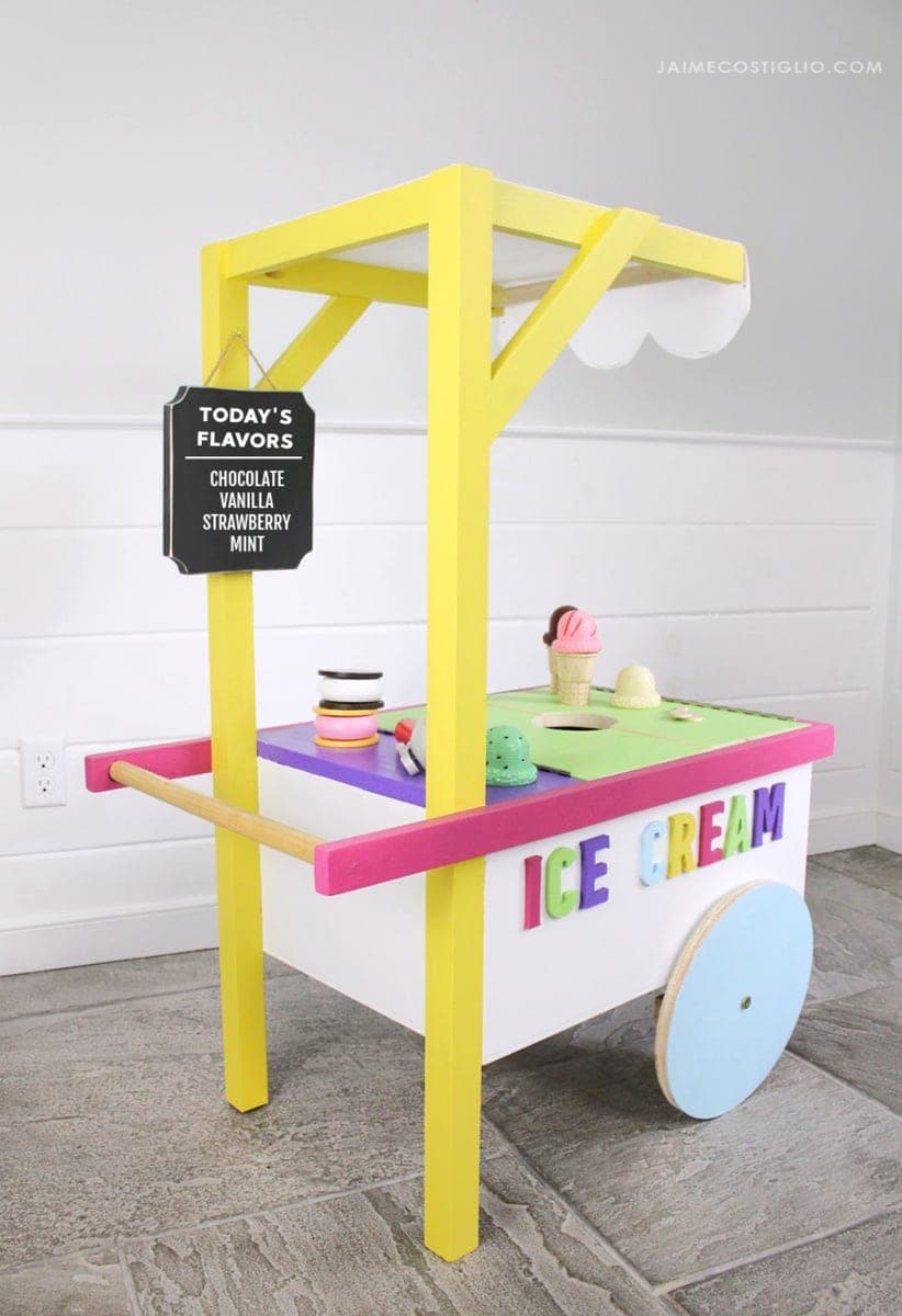 Toy wooden ice cream cart with a striped awning, wheels, and hinged serving flaps